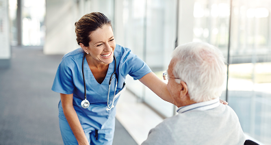 Nurse smiling at an elderly man in a wheelchair in a bright hallway.